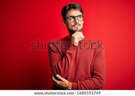 Young handsome man with beard wearing glasses and sweater standing over red background with hand on chin thinking about question, pensive expression. Smiling and thoughtful face. Doubt concept.