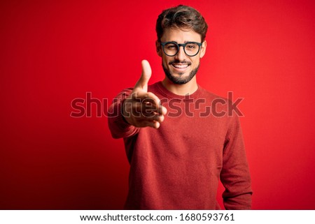 Young handsome man with beard wearing glasses and sweater standing over red background smiling friendly offering handshake as greeting and welcoming. Successful business.