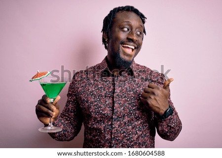 Young african american man drinking green cocktail. beverage over pink background pointing and showing with thumb up to the side with happy face smiling