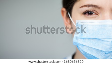 Close up of female UK EMS doctor's face,wearing blue PPE surgical protective mask,COVID-19 Coronavirus disease,global pandemic outbreak,deadly SARS-CoV-2 epidemic,grey copy space on left side of frame Royalty-Free Stock Photo #1680636820