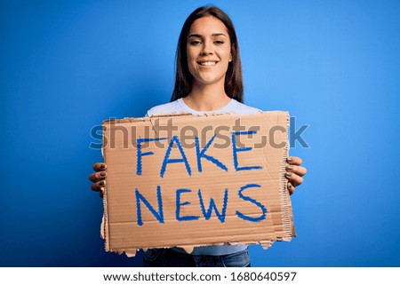 Young beautiful brunette woman holding banner with fake news message with a happy face standing and smiling with a confident smile showing teeth
