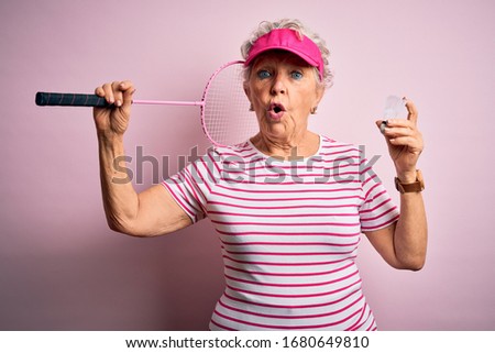 Senior beautiful sportswoman holding badminton racket over isolated pink background scared in shock with a surprise face, afraid and excited with fear expression