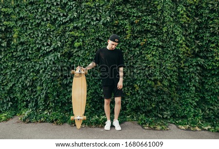 Young man in black clothes stands on a wall background with a green ivy plant, holds a longboard in his hand and looks down with a serious face.Teenager boy with a logboard on a vine background