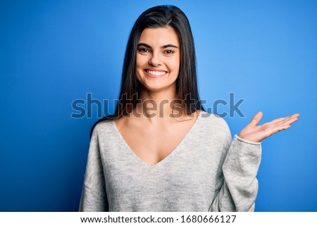 Young beautiful brunette woman wearing casual sweater standing over blue background smiling cheerful presenting and pointing with palm of hand looking at the camera.