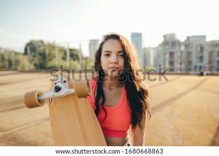 Closeup portrait of attractive hispanic girl in casual clothes standing on the street with longboard in her hands, looking into the camera with a smile on her face. Street style. Skateboard concept.