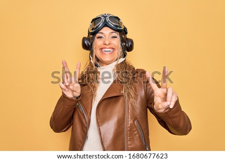 Young beautiful blonde aviator woman wearing vintage pilot helmet whit glasses and jacket smiling looking to the camera showing fingers doing victory sign. Number two.