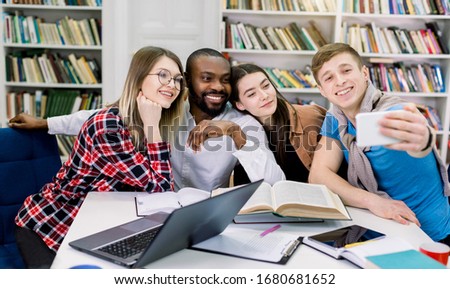 people, fun, education, technology and school concept - happy four multiethnical students making funny selfie photo in library, while studying together. Fun education, academy, school