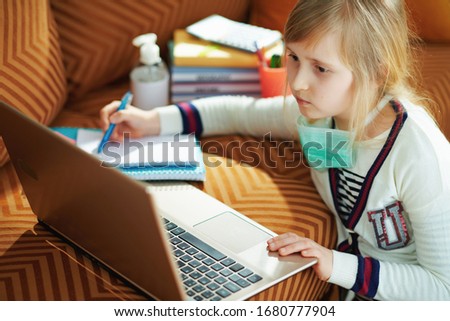 young child in white striped t-shirt with medical mask sitting near couch studying using laptop in the modern house in sunny day. Royalty-Free Stock Photo #1680777904