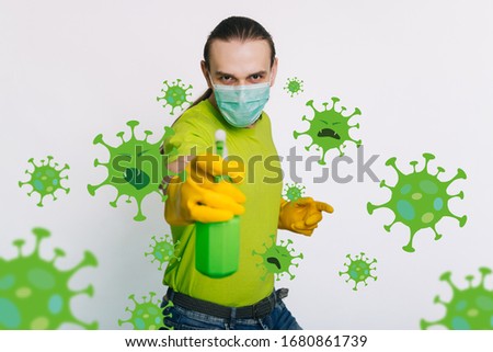 A guy in a green T-shirt and a medical mask holds in his hands a bulletizer with an antiseptic on a white background. A man kills a coronavirus. disinfects the room