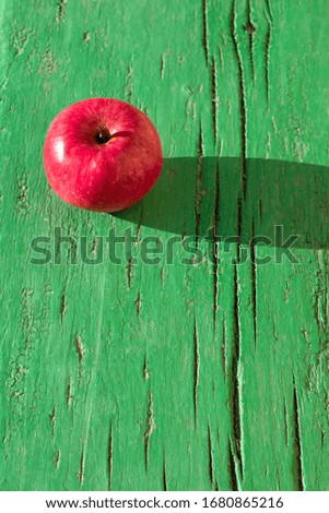 Juicy bright mature red apple on green wooden background on hard light of sunbeams with long dark shadow.
Symbol of perfection, beauty, divine gift