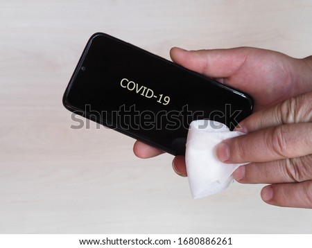 A man's hand holds a black iPhone with the inscription covid -19 and wipes it with a wet cloth on a light background, close up. Sanitizing your phone from bacteria during a coronavirus pandemic