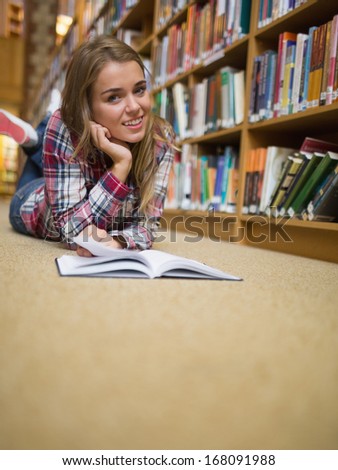 Young cheerful student lying on library floor reading book in college