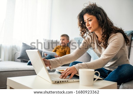 Smiling mom working at home with her child on the sofa while writing an email. Young woman working from home, while in quarantine isolation during the Covid-19 health crisis Royalty-Free Stock Photo #1680923362