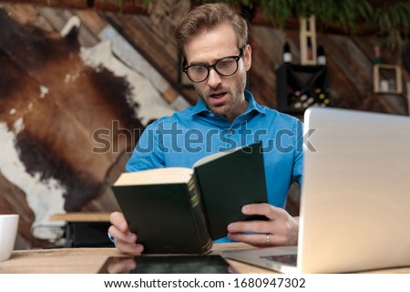 casual man wearing glasses sitting at desk and reading a book surprised at the coffeeshop