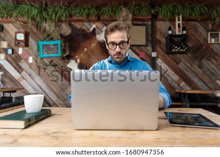 casual man wearing glasses sitting at desk and looking at laptop surprised at the coffeeshop