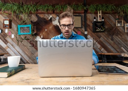 casual man wearing glasses sitting at desk and looking at his laptop shocked at the coffeeshop