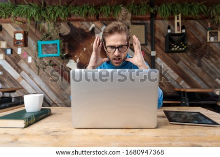 businessman wearing glasses sitting at desk and looking at his laptop fearfully surprised at the coffeeshop