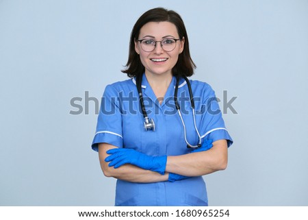 Female medical worker in blue uniform with stethoscope and gloves, confident professional woman with folded arms looking at camera, light gray background