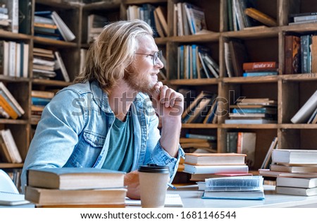 Dreamy thoughtful young teacher man male student hipster author glasses chin on hand study in library sit at desk write essay ready for exam do homework assignment smile look away education concept. Royalty-Free Stock Photo #1681146544