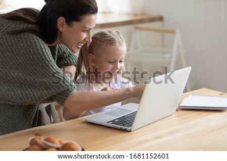 Happy young mother and cute preschooler daughter watch funny video on laptop together, smiling mom or nanny and little girl child have fun playing game on computer or studying learning online