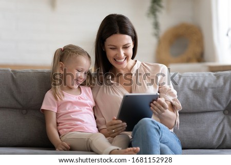 Smiling millennial nanny play with cute little girl child enjoy game on tablet relaxing on comfortable couch, happy mom and small daughter watch funny video cartoon on pad, spend weekend at home