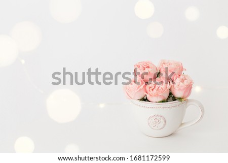 Pink peony roses in a white ceramic cup on white background with bokeh lights.