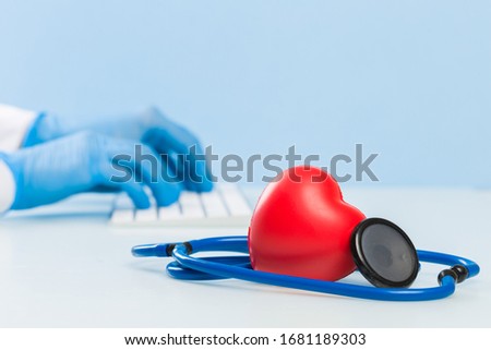 Doctor's hands in blue medical gloves are typing on a white keyboard, next to them are a dark blue stethoscope and a red heart. concept of modern medicine, cardiology