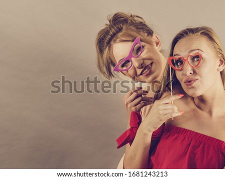 Happy two women holding paper decoration photo booth mask glasses on stick, having fun. Wedding, birthday and carnival funny accessories concept.