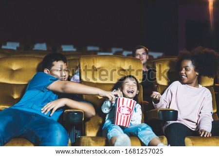 Asian kid group and african girl see movie in theater with friends. They eat a popcorn and funny cinema with big smile. Multiplex with yellow seat. 