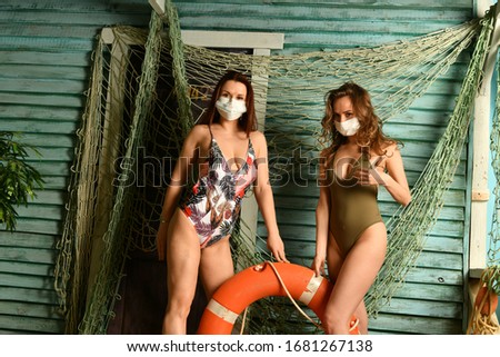 girls in bikini with a lifebuoy in medical masks on the background of a fishing house
