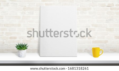 Art canvas mockup on desk leaning against the brick wall. Plant and coffee mug on desk. Scene creator with separated layers