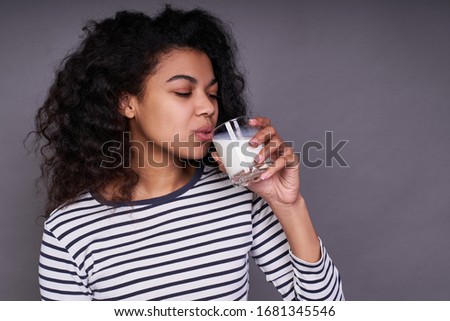 Calm cute young african american girl in a striped sweater, with curls, drinking milk, enjoying a healthy drink, posing on a gray background. The concept of calcium, vitamins.