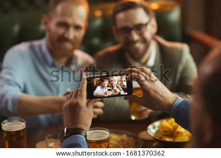 Close-up of man holding his mobile phone and taking a picture of his two friends on it while they drinking beer in cafe