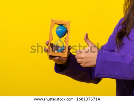 girl in a purple jacket with an hourglass in her ears on a yellow plain background. a device for counting time intervals, consisting of two transparent vessels connected by a narrow neck. hourglass.