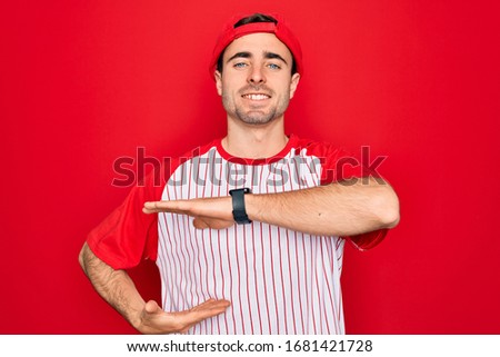 Young handsome sporty man with blue eyes wearing striped baseball t-shirt and cap gesturing with hands showing big and large size sign, measure symbol. Smiling looking at the camera. Measuring concept