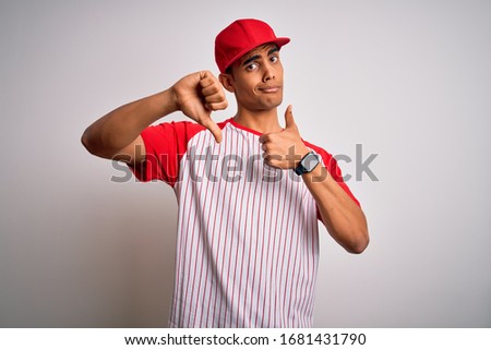 Young handsome african american sportsman wearing striped baseball t-shirt and cap Doing thumbs up and down, disagreement and agreement expression. Crazy conflict