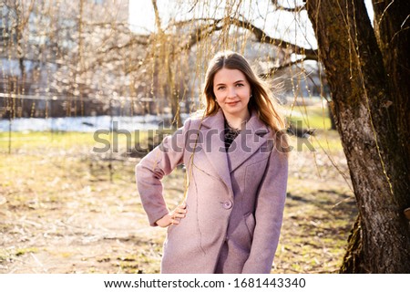 Portrait of a pretty smiling posing blonde Caucasian girl with long beautiful hair outdoors in a pink coat in a park on a wood background in the afternoon in sunny weather.
