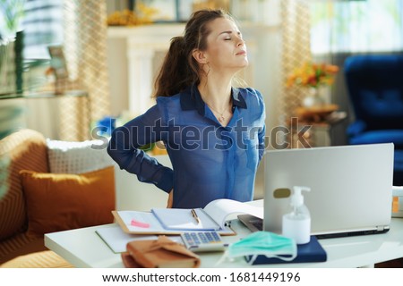 tired modern woman in blue blouse in the modern house in sunny day having back pain in temporary home office during the coronavirus epidemic. Royalty-Free Stock Photo #1681449196