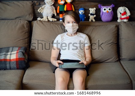 Photo in home interior of a 9 years old boy on a sofa in a medical mask with a phone tablet in his hands in quarantine