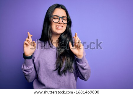 Young brunette woman wearing glasses over purple isolated background gesturing finger crossed smiling with hope and eyes closed. Luck and superstitious concept.