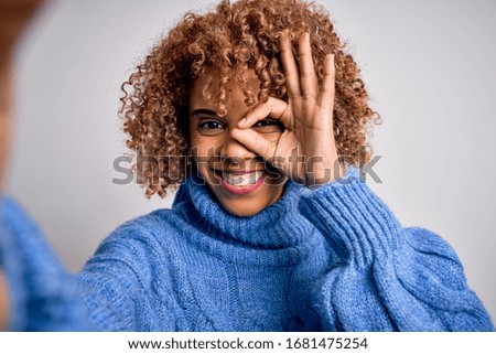 Young beautiful african american woman wearing turtleneck sweater making selfie by camera with happy face smiling doing ok sign with hand on eye looking through fingers