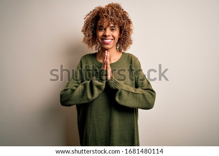 Beautiful african american woman with curly hair wearing casual sweater over white background praying with hands together asking for forgiveness smiling confident.