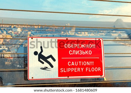 Text on sign: Caution! Slippery floor. Rectangular red and white plate with white text and silhouette of a falling man. Blurred cityscape in the background. New Pedestrian Bridge, Kyiv, Ukraine.
