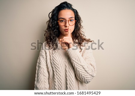 Beautiful woman with curly hair wearing casual sweater and glasses over white background asking to be quiet with finger on lips. Silence and secret concept.