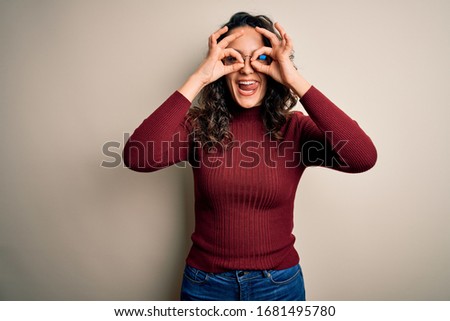 Beautiful woman with curly hair wearing casual sweater and glasses over white background doing ok gesture like binoculars sticking tongue out, eyes looking through fingers. Crazy expression.