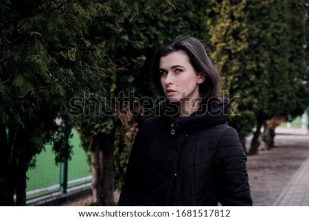 Portrait of a young pretty teen girl in a coat between thuja on the street in autumn evening.