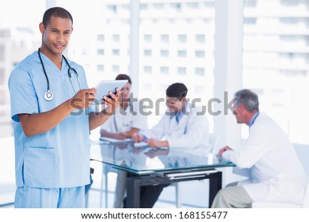 Male surgeon using digital tablet with group around table in background at hospital