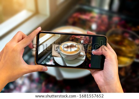 Hand holding mobile phone and take a photo Coffee cup on blurred coffee shop background with sunlight.