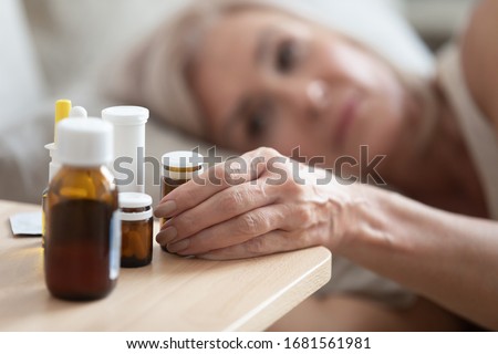 Close up focus on wrinkled female hand taking glass bottle with pills. Head shot unhealthy middle aged woman lying in bed, feeling unwell, taking medicine from flu or insomnia, suffering from cold. Royalty-Free Stock Photo #1681561981