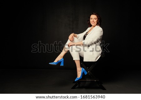 Happy business woman in a white jacket sits on a chair and looks at the camera over black background. Free space.